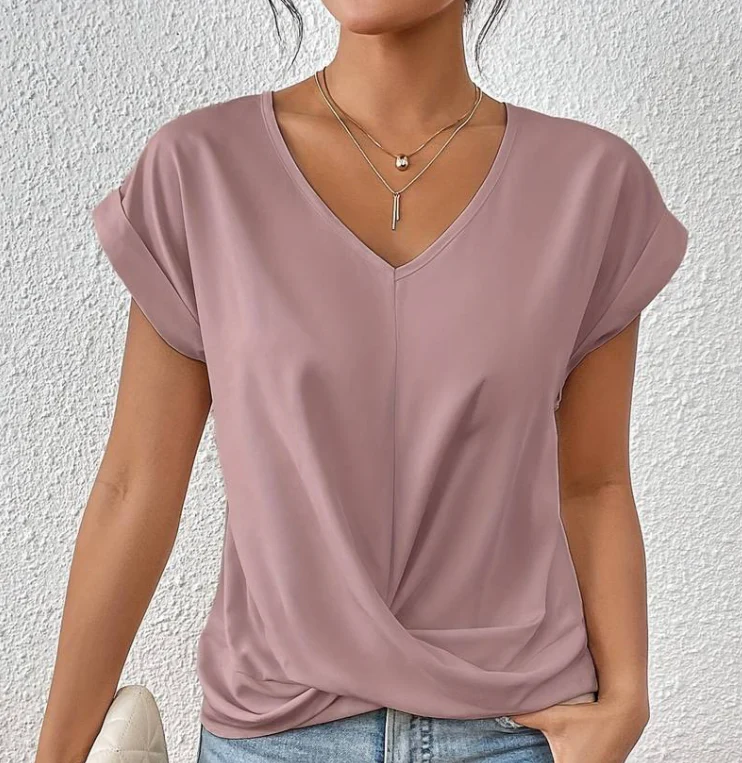 Femme portant un t-shirt rose à col en V, manches courtes, avec collier doré, posant devant un mur blanc. Mode féminine décontractée, style chic.