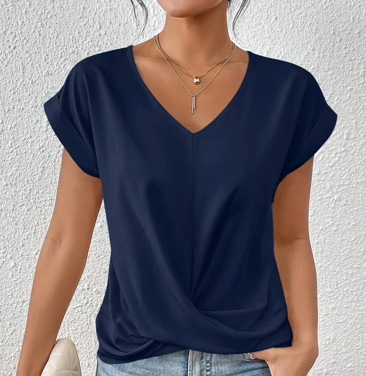 Femme portant un t-shirt bleu marine à col en V, manches courtes, avec colliers dorés superposés, posant contre un mur blanc texturé.