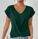 Femme portant un t-shirt vert foncé à col en V, manches courtes, avec colliers dorés superposés, devant un mur blanc texturé.