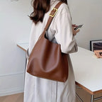 Femme portant un sac à main en cuir marron, style épaule, avec boucle dorée, sur fond de bureau moderne. Mode féminine, accessoires élégants.