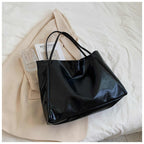 Sac à main noir en cuir brillant sur un manteau beige, posé sur un lit blanc avec un livre et un verre transparent. Accessoire mode élégant.