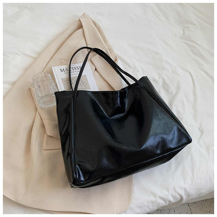 Sac à main noir en cuir brillant sur un manteau beige, posé sur un lit blanc avec un livre et un verre transparent. Accessoire mode élégant.