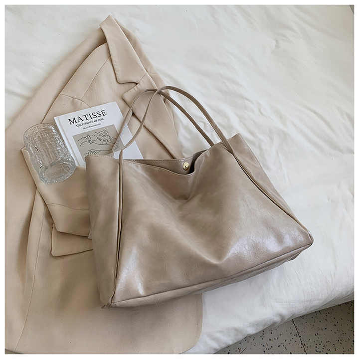Sac à main en cuir beige posé sur un manteau crème, avec un livre "Matisse" et un verre en cristal, sur un lit blanc. Accessoires élégants et modernes.