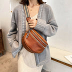 Femme portant un cardigan gris et une robe beige, tenant un sac banane en cuir marron avec bandoulière rayée. Mode automne, accessoires tendance.