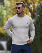 Homme portant un pull torsadé beige, lunettes de soleil, debout à l'extérieur avec un fond flou de verdure automnale. Mode masculine élégante.