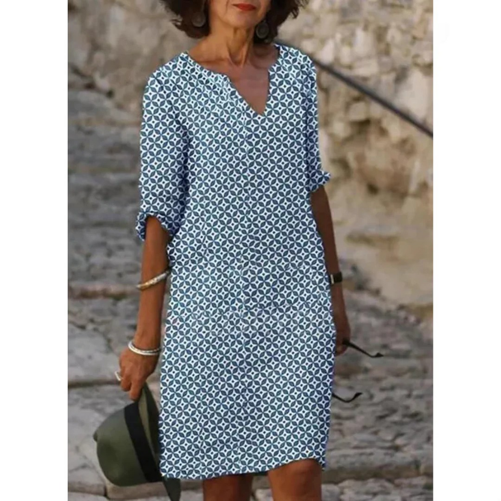 Femme portant une robe d'été bleue à motifs géométriques, manches trois-quarts, marchant sur un chemin pavé, tenant un chapeau vert.