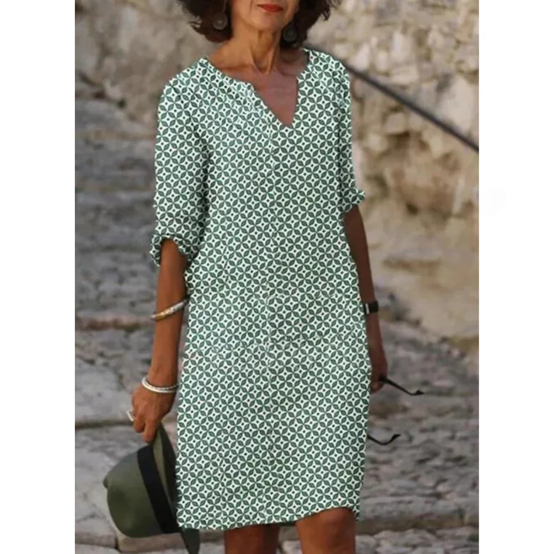 Femme portant une robe à motifs géométriques verts et blancs, manches trois-quarts, tenant un chapeau vert, marchant sur un chemin pavé.