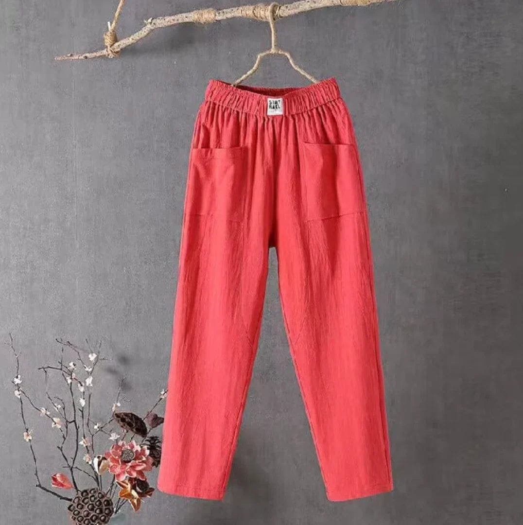 Pantalon rouge en lin suspendu sur une branche, style décontracté, poches avant, fond gris, décoration florale en bas à gauche.
