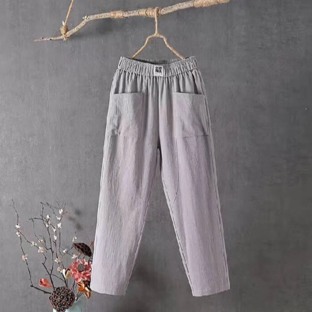 Pantalon gris en lin suspendu à une branche, fond gris, décor floral en bas à gauche. Mode éco-responsable, style décontracté, tendance minimaliste.