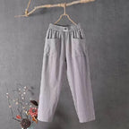 Pantalon gris en lin suspendu à une branche, fond gris, décor floral en bas à gauche. Mode éco-responsable, style décontracté, tendance minimaliste.