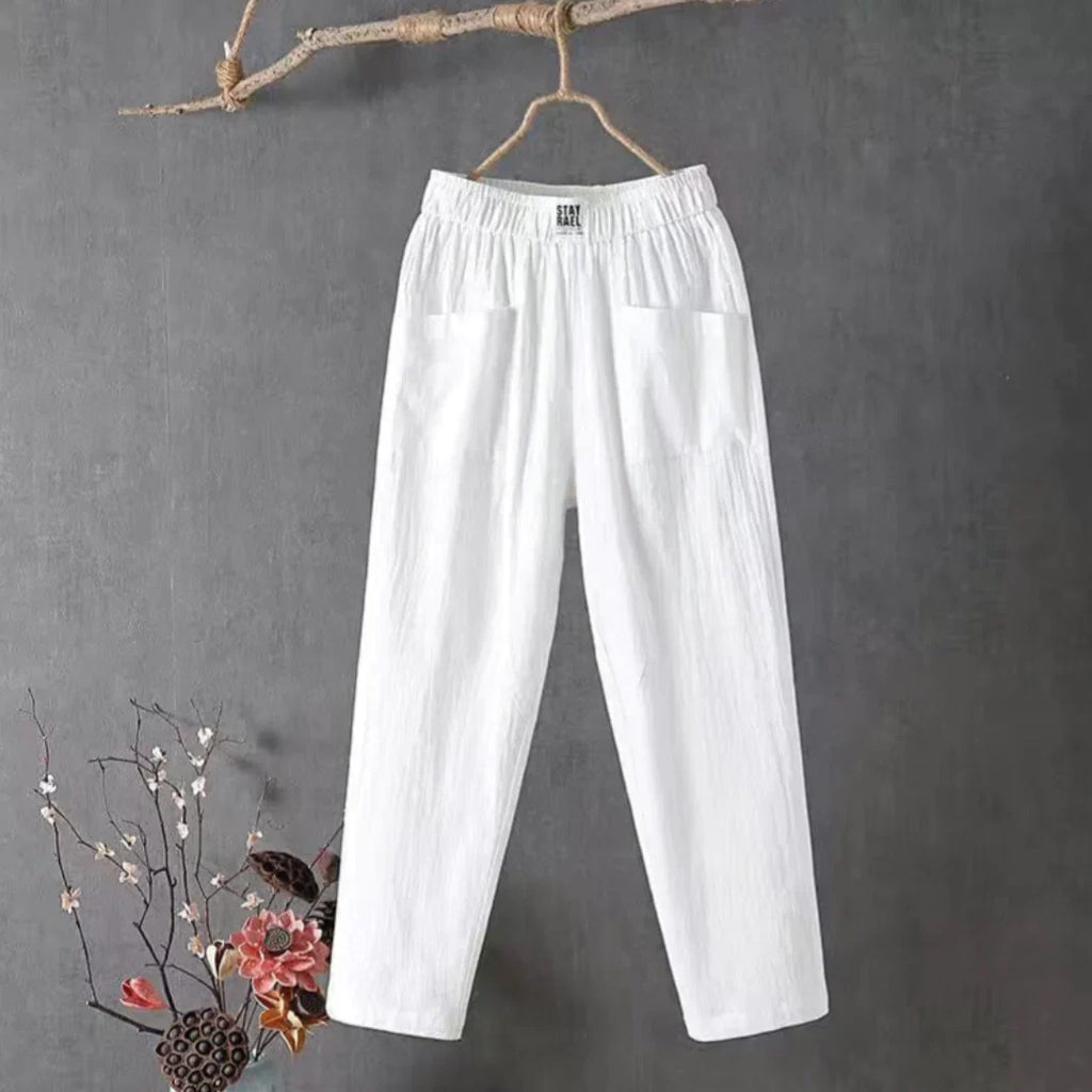 Pantalon blanc en lin suspendu sur une branche, fond gris, décor floral en bas à gauche. Mode éthique, vêtements légers, style décontracté.