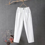 Pantalon blanc en lin suspendu sur une branche, fond gris, décor floral en bas à gauche. Mode éthique, vêtements légers, style décontracté.