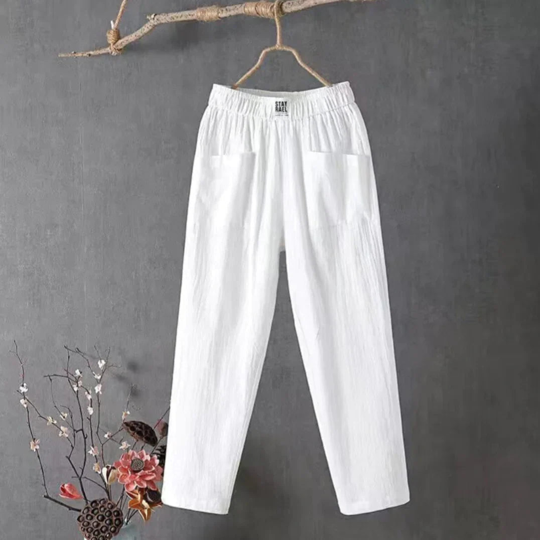 Pantalon blanc en lin suspendu sur une branche, fond gris, décor floral en bas à gauche. Mode éthique, vêtements légers, style décontracté.