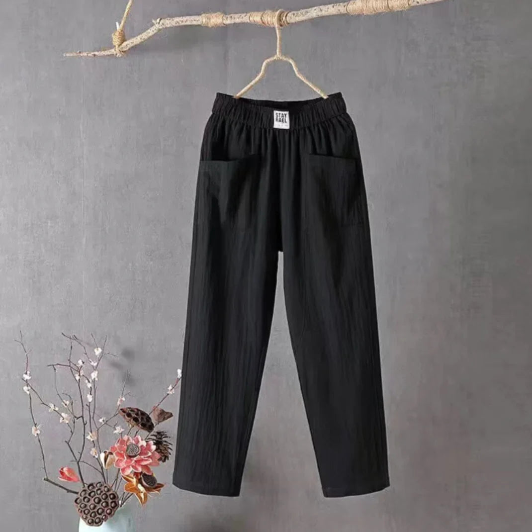 Pantalon noir en lin suspendu sur une branche, fond gris, décor floral en bas à gauche. Mode minimaliste, style décontracté, vêtements en lin.