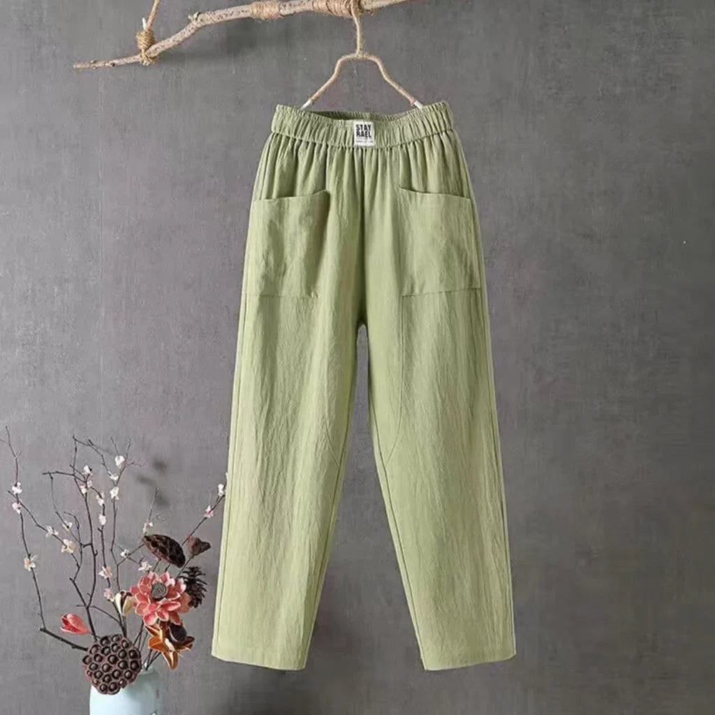 Pantalon vert en lin suspendu sur un cintre, avec poches avant, sur fond gris. Décoration florale en vase bleu à gauche. Mode éco-responsable.