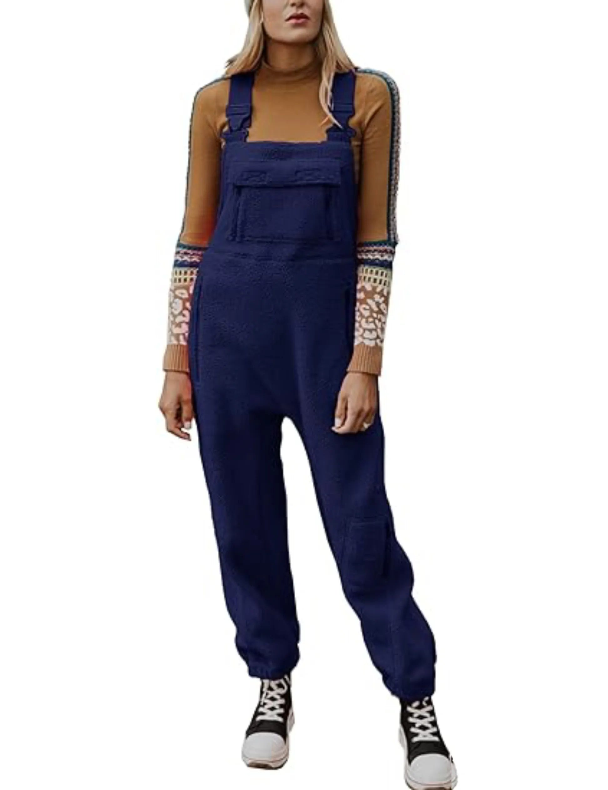 Femme portant une salopette bleue en laine, pull à manches longues marron avec motifs, baskets noires et blanches. Mode automne-hiver tendance.
