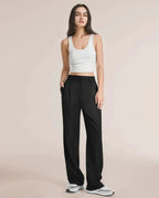 Femme portant un débardeur blanc et un pantalon noir ample, posant debout sur un fond neutre. Mode décontractée, élégance minimaliste.