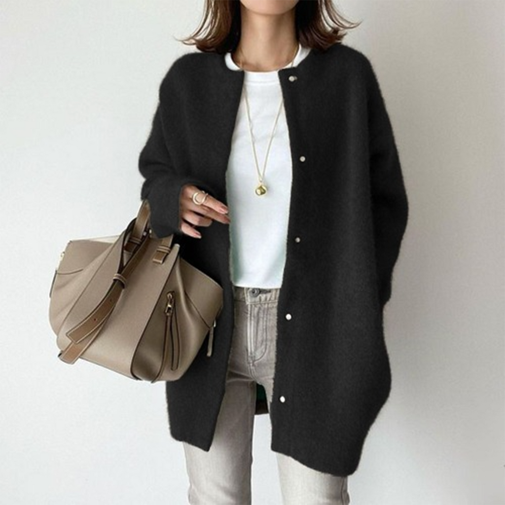 Femme portant un long cardigan noir, t-shirt blanc, jean gris, collier doré, tenant un sac à main beige. Mode automne-hiver élégante et décontractée.