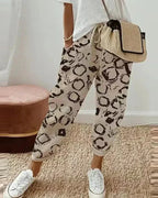 Femme portant un pantalon beige à motifs noirs, t-shirt blanc, sac en paille, baskets blanches. Mode décontractée, tendance, style urbain.