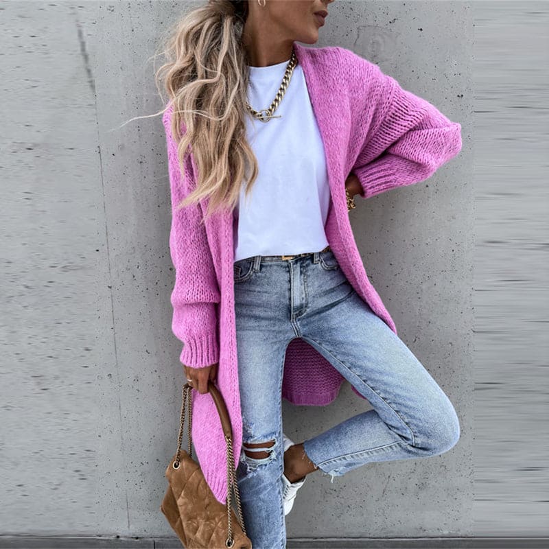 Femme portant un cardigan rose, t-shirt blanc, jeans déchirés, sac marron, posant contre un mur gris. Mode décontractée, style tendance.