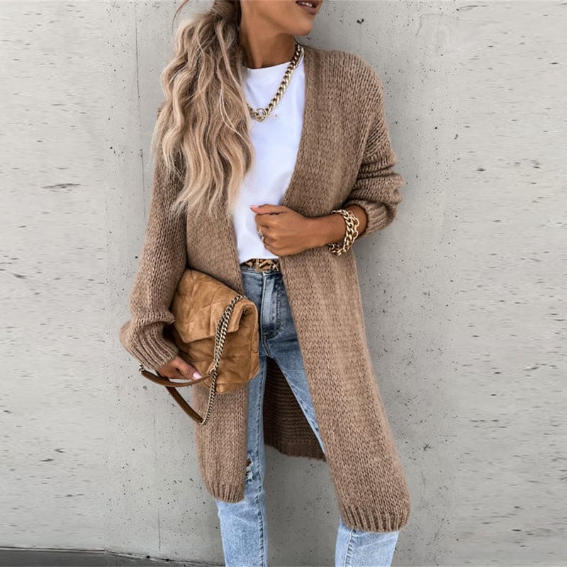 Femme portant un cardigan long beige, t-shirt blanc, jean délavé, sac à main camel, accessoires dorés, posant contre un mur gris.