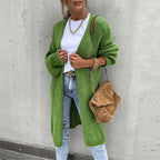 Femme portant un cardigan vert, t-shirt blanc, jeans déchirés, sac à main marron, chaîne dorée, posant contre un mur gris. Mode automne chic.