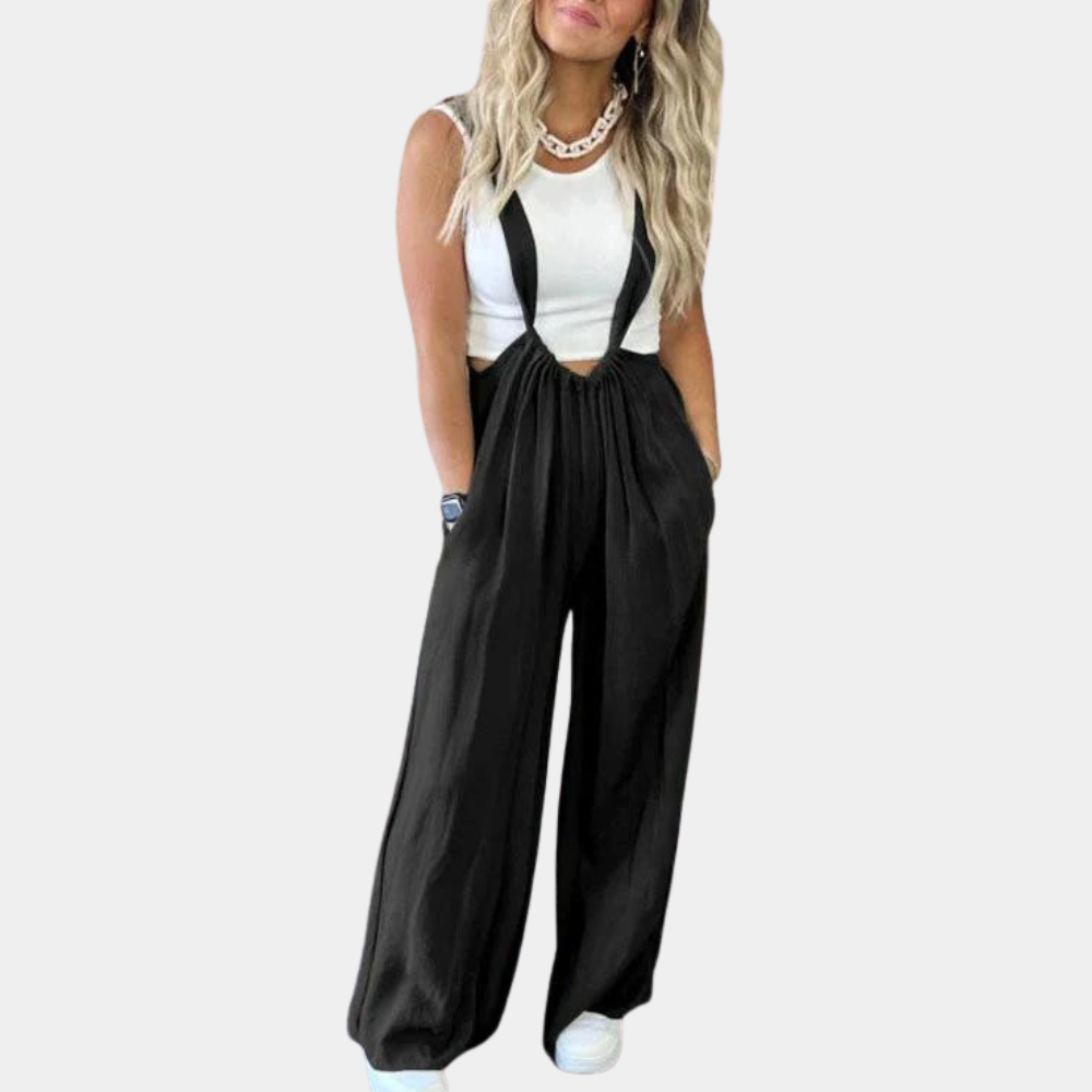 Femme portant une salopette noire élégante avec un haut blanc sans manches, collier doré, cheveux blonds ondulés, et baskets blanches.