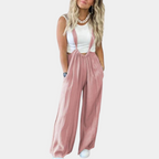 Femme portant une salopette rose ample avec un haut blanc sans manches, cheveux blonds ondulés, posant sur fond blanc. Mode décontractée chic.
