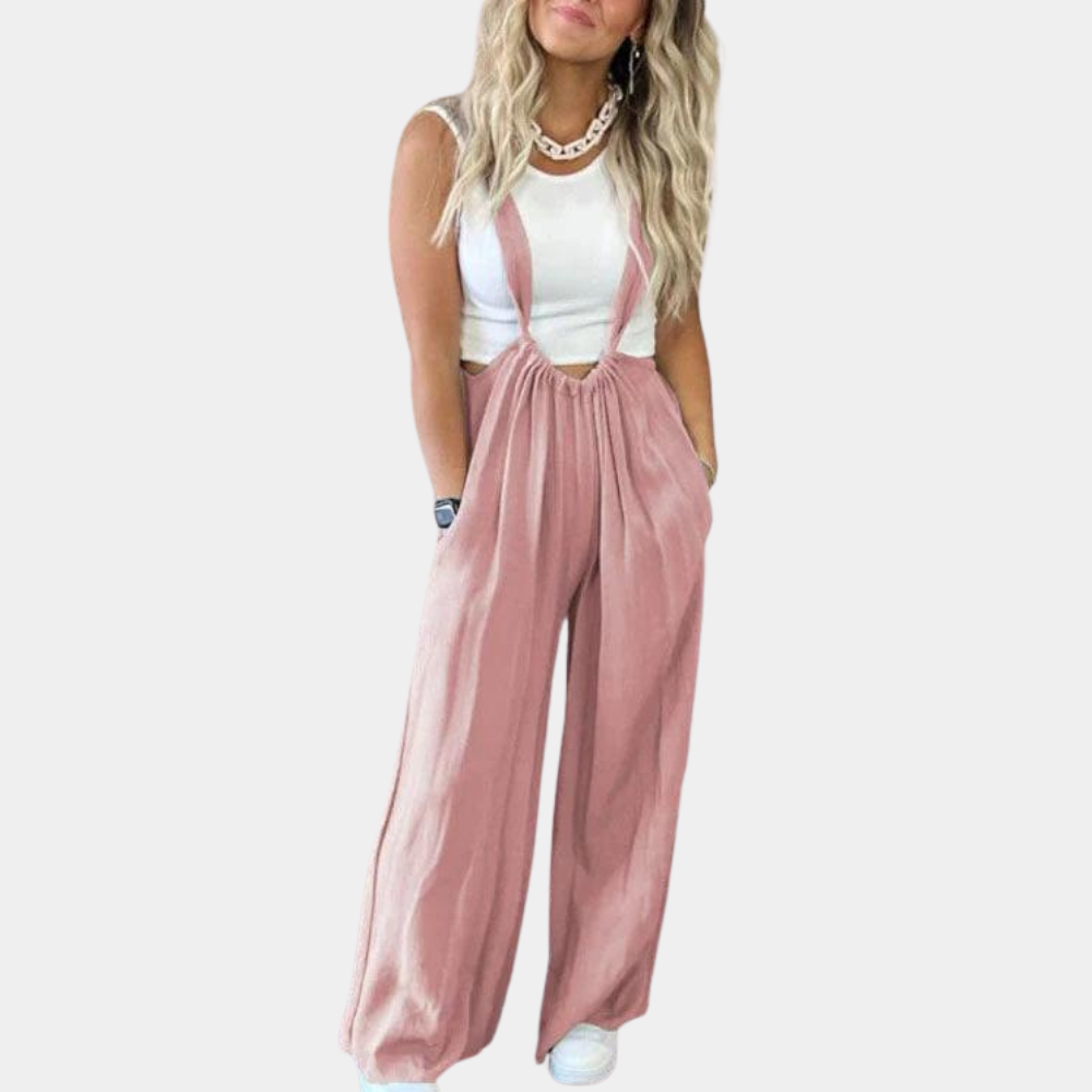 Femme portant une salopette rose ample avec un haut blanc sans manches, cheveux blonds ondulés, posant sur fond blanc. Mode décontractée chic.