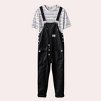 Salopette noire en denim avec boutons, portée sur un t-shirt rayé blanc et noir. Mode décontractée, tendance, vêtements pour homme.
