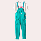 Salopette turquoise en coton avec boutons argentés, poches avant, portée sur un t-shirt blanc imprimé rose. Mode décontractée, tendance urbaine.