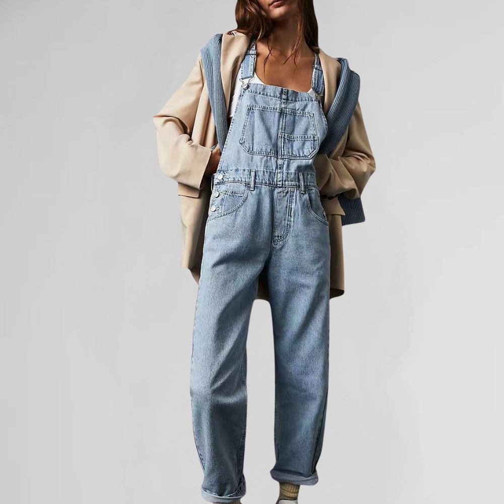 Femme portant une salopette en jean bleu clair, associée à un pull gris et un manteau beige. Mode décontractée, tendance automne-hiver.