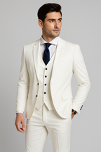 Homme en costume blanc élégant, cravate noire, chemise blanche, posant sur fond gris. Mode masculine, tenue formelle, style sophistiqué.
