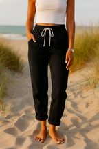 Femme en débardeur blanc et pantalon noir sur plage, pieds nus dans le sable, herbes maritimes en arrière-plan, montre-bracelet dorée visible.