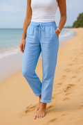 Femme en pantalon bleu clair sur plage de sable, portant un débardeur blanc. Mode estivale, détente bord de mer, style décontracté.