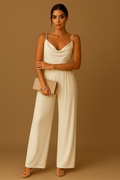 Femme en combinaison beige élégante, tenant une pochette assortie, posant sur fond marron. Mode féminine chic, tenue soirée tendance.