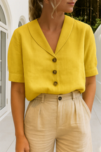 Femme portant une chemise jaune à col large et boutons marron, associée à un pantalon beige, devant une maison moderne. Mode estivale élégante.