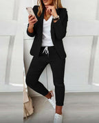 Femme en tenue décontractée chic, portant un blazer noir, t-shirt blanc, pantalon noir et baskets blanches, tenant un smartphone.