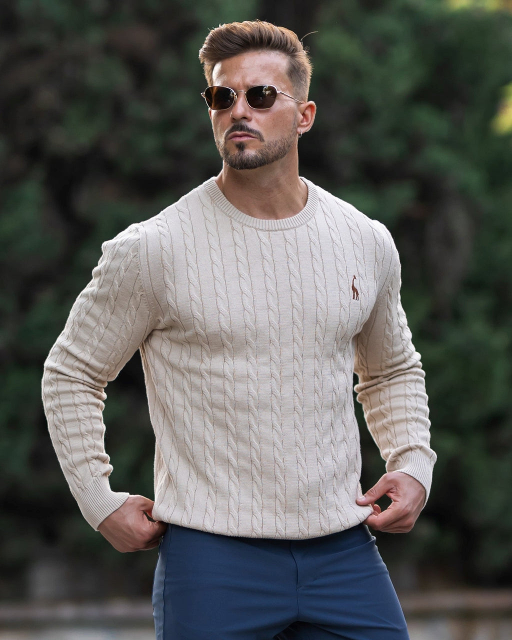 Homme portant un pull torsadé beige et des lunettes de soleil, posant à l'extérieur devant un fond flou de verdure. Mode masculine élégante.