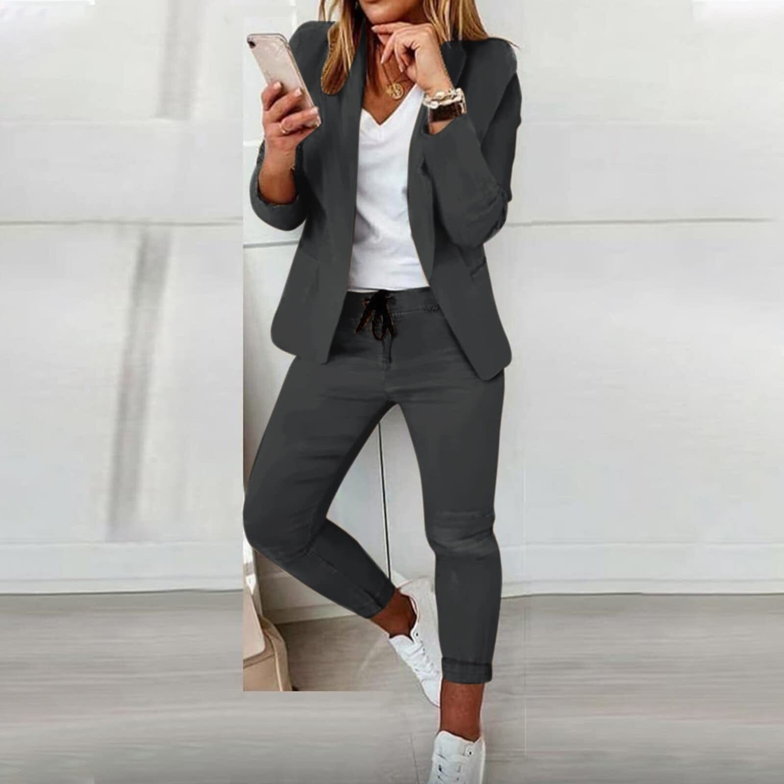 Femme en costume gris moderne, tenant un smartphone, portant des baskets blanches. Mode professionnelle décontractée, tendance et élégante.