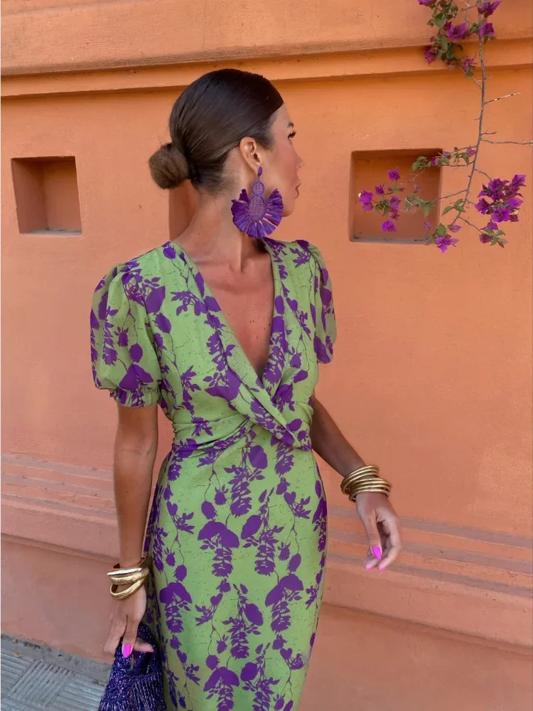 Femme en robe verte à motifs violets, accessoires dorés, boucles d'oreilles violettes, mur orange en arrière-plan, fleurs violettes.