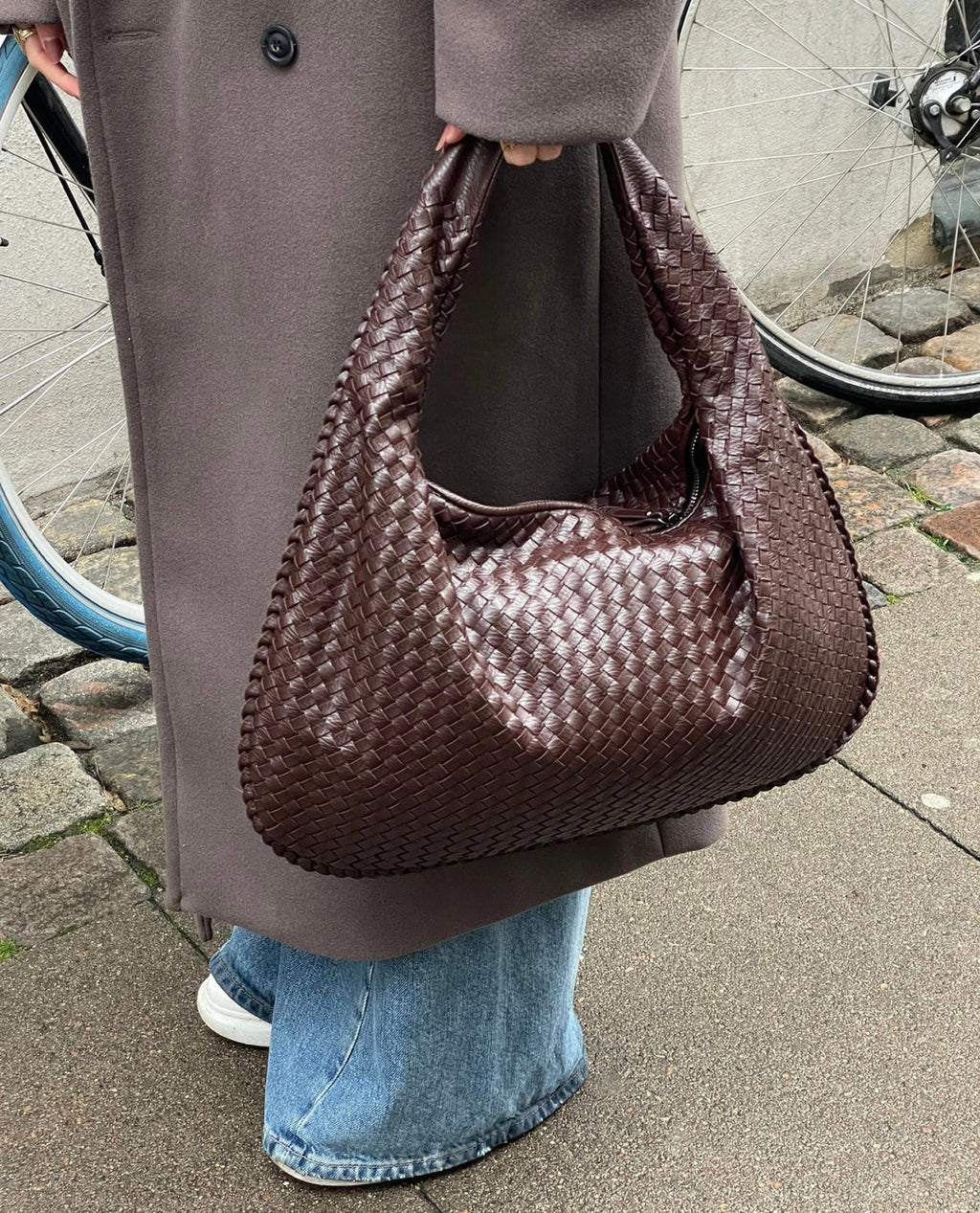 Sac à main en cuir tressé marron porté par une personne en manteau gris et jean, avec une roue de vélo en arrière-plan sur un trottoir urbain.