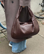 Sac à main en cuir tressé marron porté par une personne en manteau gris et jean, avec une roue de vélo en arrière-plan sur un trottoir urbain.