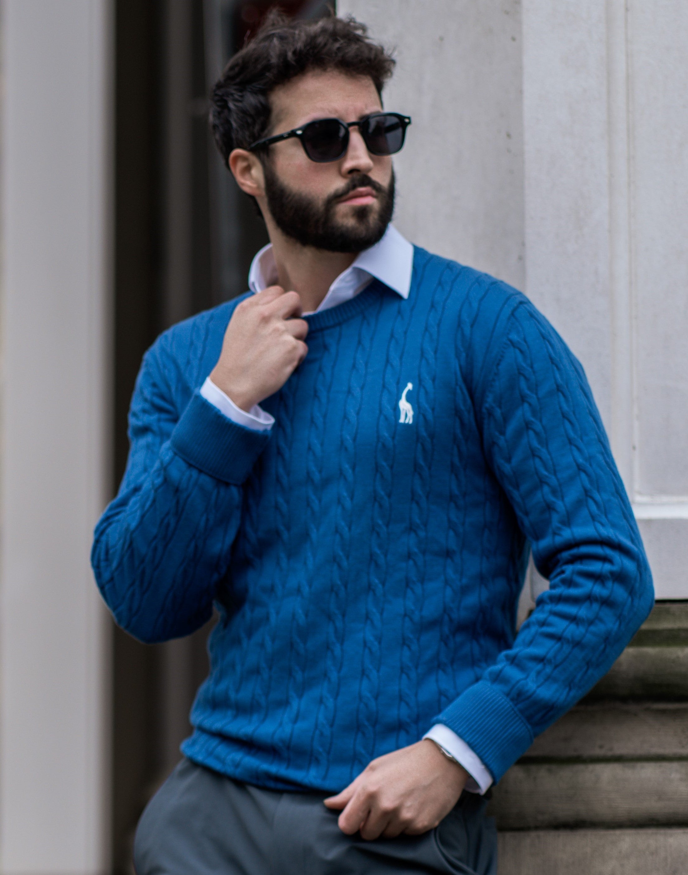 Homme portant un pull bleu en maille torsadée, chemise blanche et lunettes de soleil noires, posant devant un mur blanc. Mode masculine élégante.