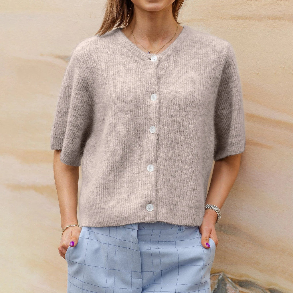 Femme portant un cardigan beige en laine avec boutons blancs, manches courtes, associé à un pantalon bleu à carreaux. Mode automne chic et décontractée.