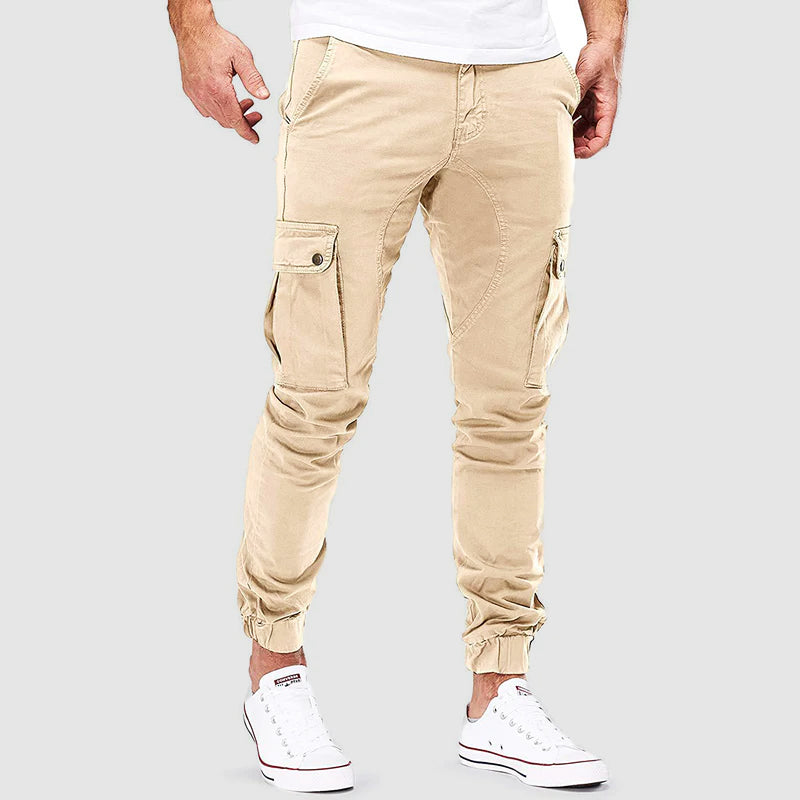 Pantalon cargo beige pour homme, style décontracté, poches latérales, coupe ajustée, porté avec des baskets blanches, fond gris.