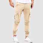 Pantalon cargo beige pour homme, style décontracté, poches latérales, coupe ajustée, porté avec des baskets blanches, fond gris.