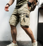 Homme portant un short cargo beige, montre noire et baskets blanches, posant contre un mur blanc. Mode décontractée, style urbain, été.