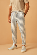 Homme portant un pantalon à rayures verticales blanches et noires, avec un t-shirt beige et des baskets blanches, sur fond beige.