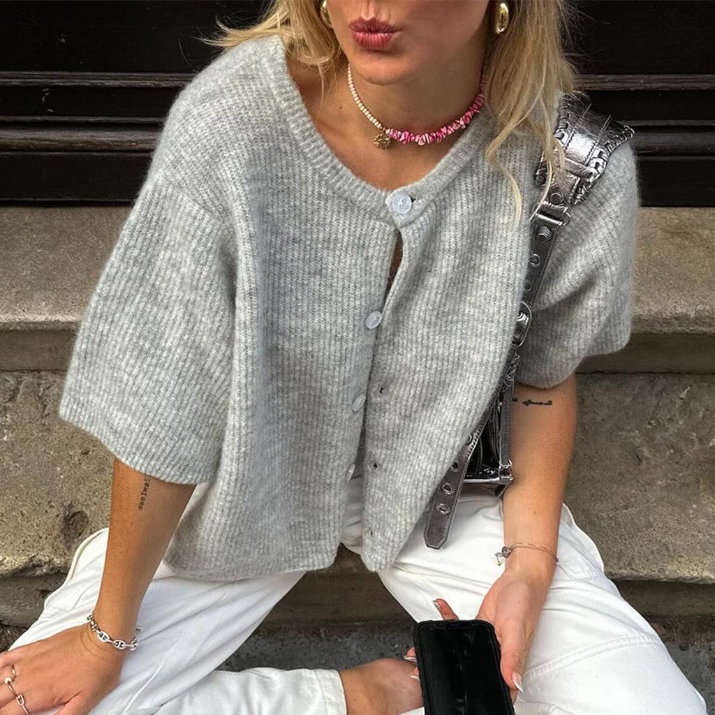 Femme portant un cardigan gris en tricot, collier rose, pantalon blanc, assise sur un trottoir. Mode décontractée, accessoires tendance.